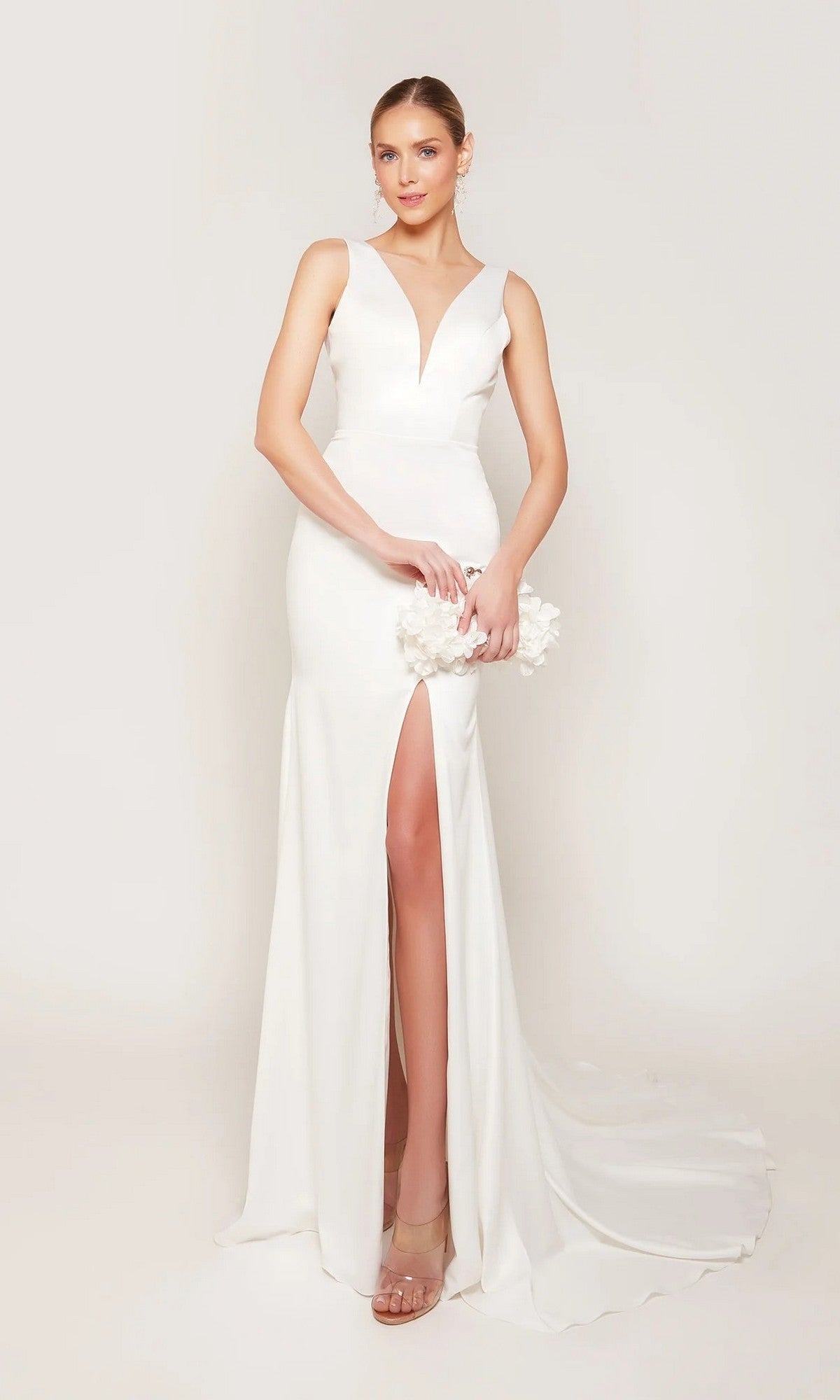 Wearing the Alyce 7091 long white dress with a sleeveless plunging V-neck, thigh-high slit, and train, a woman holds a white floral clutch against a plain light background.